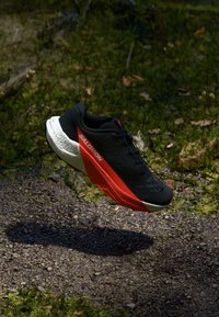Crna tenisica za trčanje s crvenim naglaskom sa strane, laganog dizajna, teksturiranim potplatom i mrežastim gornjim dijelom, postavljena iznad prirodne, šljunčane površine.