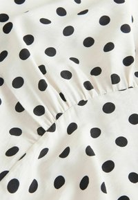 Balts audums ar melniem punktiņiem, kuram ir savilkta elastīga detaļa, radot nelielas locījumu kroku un tekstūru.