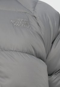 Grijze pufferjack met een gequilte ontwerp. Het North Face-logo is in grijs op de voorkant geborduurd. Glad, glanzend oppervlak.