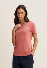 Camiseta de algodón color rosa con mangas cortas, que presenta un diseño "FY" adornado con cristales en el lado izquierdo del pecho. Textura suave, corte relajado.