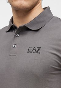 Grå polo shirt med knaplukket krave, med en sort broderet logo på venstre bryst. Fremstillet af en glat, tekstureret stof.