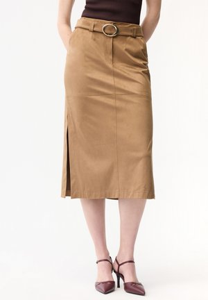 Beigefarbener Rock knielang mit Gürtel und Schlitz auf der linken Seite, kombiniert mit dunkelroten spitz zulaufenden Absatzschuhen und einem dunklen ärmellosen Oberteil.