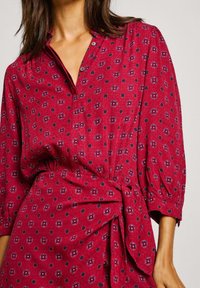 Vestido rojo de botón con cintura fruncida, que presenta un estampado floral y de lunares. Tejido texturizado y mangas abullonadas.