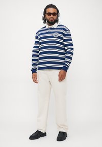 Mørkeblå og hvite stripete langermet polo skjorte med krage og emblem, kombinert med beige bukser og svarte slip-on sko.