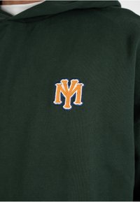 Sweat à capuche vert foncé avec un logo NY stylisé brodé en orange et blanc sur la poitrine.