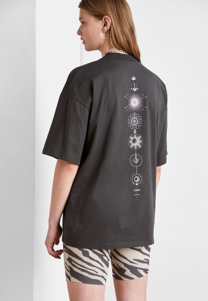 Tmavě šedé oversized tričko s bílými a růžovými geometrickými vzory na zadní straně. Spárováno s béžovými šortkami se zebrovaným vzorem.