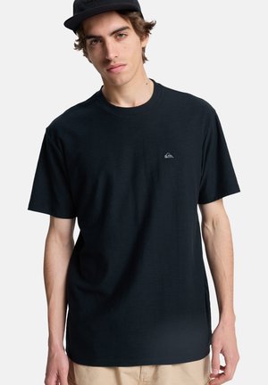 Junger Mann mit schwarzer Mütze, schwarzem T-Shirt mit kleinem Logo auf der Brust und beiger Hose, steht vor einem einfarbigen hellen Hintergrund.