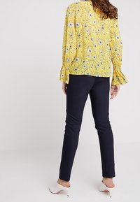 Blouse florale jaune à manches longues et poignets à volants, associée à un pantalon noir ajusté et des chaussures à talons blancs. Vue de dos présentée.