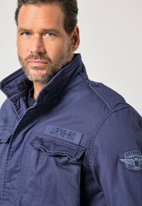 Mies, jolla on lyhyet tummat hiukset ja parta, pukeutunut siniseen sotilastyyliseen takkiin, jossa on merkkejä ja pystytetty kaulus, katsoo suoraan eteenpäin.