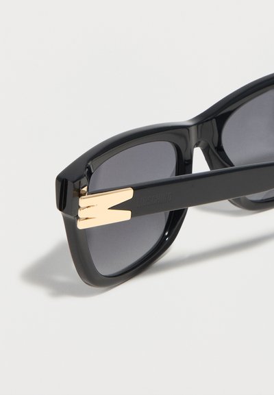 Μαύρα ορθογώνια γυαλιά ηλίου με σκουρόχρωμους φακούς και χρυσή λεπτομέρεια "M" στη σύνδεση του βραχίονα, σε ανοιχτό φόντο.