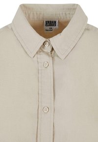 Beige skjorte med krage og knappelukking, som har fire knapper og en etikett som sier "URBAN CLASSICS" inni kragen.