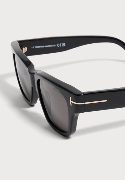 Lunettes de soleil rectangulaires noires avec verres teintés et détail doré en forme de T sur les branches, vue en angle montrant le nom de la marque et la charnière.