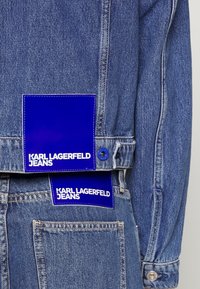 Džínová bunda má světle modrý odstín s viditelným šitím. Má dvě modré textilní cedulky s bílým nápisem „KARL LAGERFELD JEANS".