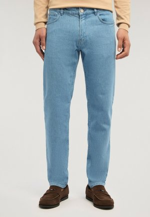 Mann trägt hellblaue Jeans mit geradem Schnitt, beigefarbenen Pullover und braune Wildleder-Loafer, steht vor einfachem hellem Hintergrund.