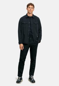 Camicia button-up blu scuro con due tasche sul petto, abbinata a pantaloni neri e sneaker con motivo. Design semplice con una vestibilità ampia.