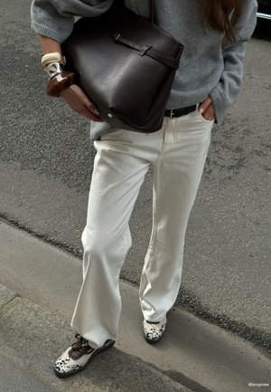 Persoon draagt witte broek, grijze trui, sneakers met patroon, houdt een grote donkerbruine leren tas vast en draagt opvallende armbanden.