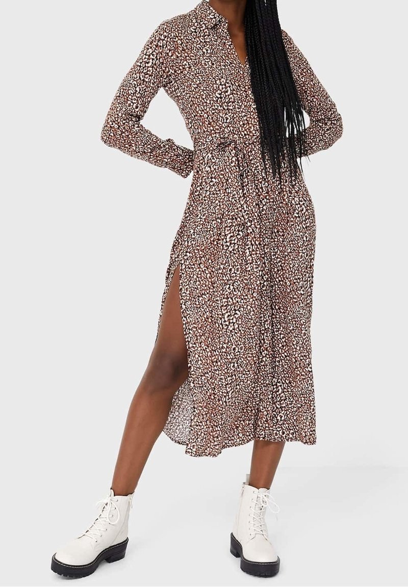 Femme portant une robe midi à manches longues à imprimé léopard avec une fente latérale, associée à des bottines blanches à lacets, debout les mains sur les hanches.