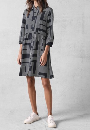 Femme portant une robe-chemise à manches longues avec un motif géométrique noir et blanc et des baskets blanches, debout contre un mur gris texturé.