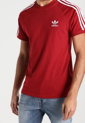T-shirt en coton rouge avec logo Adidas blanc sur la poitrine, présentant des accents rayés blancs sur les épaules et un col rond.