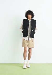 Schwarze ärmeloses Jacket über einem beigen Hemd, kombiniert mit khakifarbenen Shorts und weißen Sneakers. Schlichte Gestaltung ohne sichtbare Muster oder Akzente.