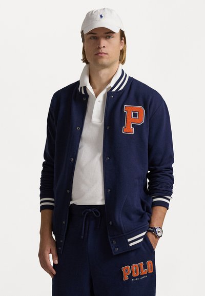 Navy blue varsity jacket with striped cuffs, "P" appliqué in orange, worn over a white polo shirt, paired with matching navy pants.