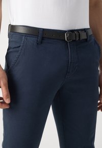 Tmavě modré chinos kalhoty s hladkým povrchem, vybavené bočními kapsami a černým koženým opaskem s jednoduchou stříbrnou přezkou. Zapínání na knoflíky v pase.