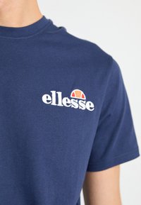 T-shirt en coton bleu marine avec un logo brodé blanc et un demi-cercle orange au-dessus de la lettre "e" sur la partie gauche de la poitrine.