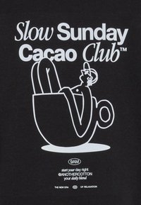 T-shirt nera con grafica bianca che riporta il testo "Slow Sunday Cacao Club" e un'illustrazione di una persona sdraiata in una grande tazza di caffè.