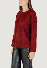 Maglione a coste bordeaux con orlo a coste e dettagli con bottoni sui lati. Presenta maniche lunghe e una vestibilità rilassata, abbinato a pantaloni in pelle nera.