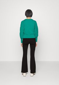 Teal groene trui met getextureerde pofmouwen, aansluitende ribbelzoom, gecombineerd met zwarte flared broeken en crèmekleurige enkellaarsjes. Effen achtergrond.