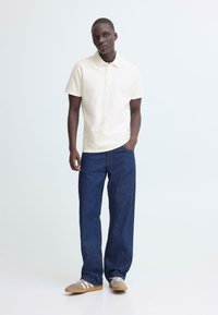 Beige poloshirt met korte mouwen, donkerblauwe wijde jeans, en grijze sneakers met witte accenten. Effen texturen, neutraal kleurenpalet.
