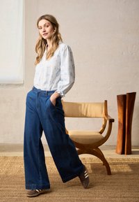 Femme aux longs cheveux blonds portant un chemisier blanc à fleurs, un jean bleu à jambes larges et des baskets marron, debout à côté d'une chaise en bois.
