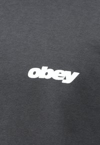 Sweatshirt cinza escuro com uma textura suave, apresentando um logotipo branco "obey" centralizado no peito. Design simples, sem padrões ou acentos adicionais.