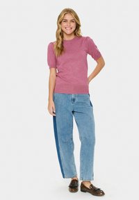 Lächelnde Frau steht mit der Hand auf der Hüfte, trägt ein kurzärmliges lila Oberteil, zweifarbige blaue Jeans und braune Slipper vor einem weißen Hintergrund.