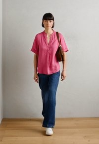 Camicia di lino rosa a maniche corte con scollo a V, abbinata a jeans a campana blu e sneaker bianche. Una borsa a tracolla marrone completa il look.