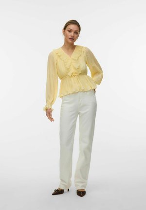 Kvinne står iført en gul bluse med volanger og lange ermer, hvite bukser og mørke spisse sko med lave hæler.