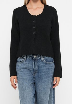 Femme portant un cardigan noir à manches longues boutonné et un jean taille haute bleu, debout devant un fond blanc uni.