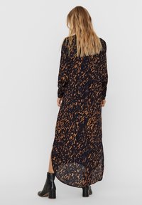 Mujer con cabello rubio largo que lleva un vestido largo de estampado negro y marrón y botas negras hasta el tobillo, de pie frente a un fondo blanco liso.