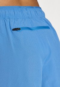 Blå treningsshorts laget av lett stoff med perforert tekstur. Har en innsatt glidelåslomme i en kontrasterende blå farge.