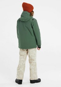 Veste verte imperméable avec capuche, manches élastiques. Pantalons beiges avec un motif graffiti subtil. Bottes de neige noires complètent le look.