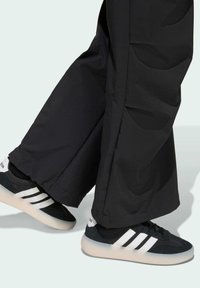 Black wide-leg pants paired with black sneakers featuring three white stripes, suede and mesh material, and a light-colored rubber sole.