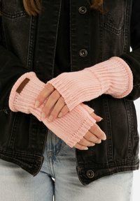 Gants sans doigts en tricot rose clair avec texture côtelée et poignet. Détail d'étiquette marron au niveau du poignet. Portés par-dessus une veste en denim noire.