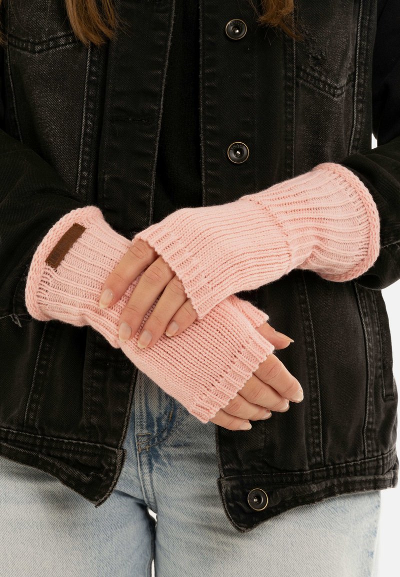 Gants sans doigts en tricot rose clair avec texture côtelée et poignet. Détail d'étiquette marron au niveau du poignet. Portés par-dessus une veste en denim noire.