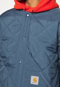 Niebieska pikowana kurtka na zatrzaski noszona na czerwonym polarze z kapturem, z naszywką z logo Carhartt na dolnej przedniej kieszeni.