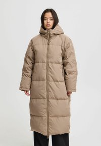 Lungo cappotto imbottito beige con cappuccio, dotato di zip frontale, maniche ampie e motivo trapuntato. Finitura liscia e testurizzata.