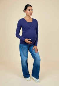 Top de manga larga azul marino con un diseño cruzado y detalle fruncido, combinado con pantalones vaqueros de pierna ancha en azul claro y zapatillas blancas.