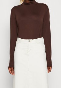 Brun långärmad topp med hög hals som är kombinerad med en vit denimkjol, som har synlig sömnad och en framknäppning med knappar.