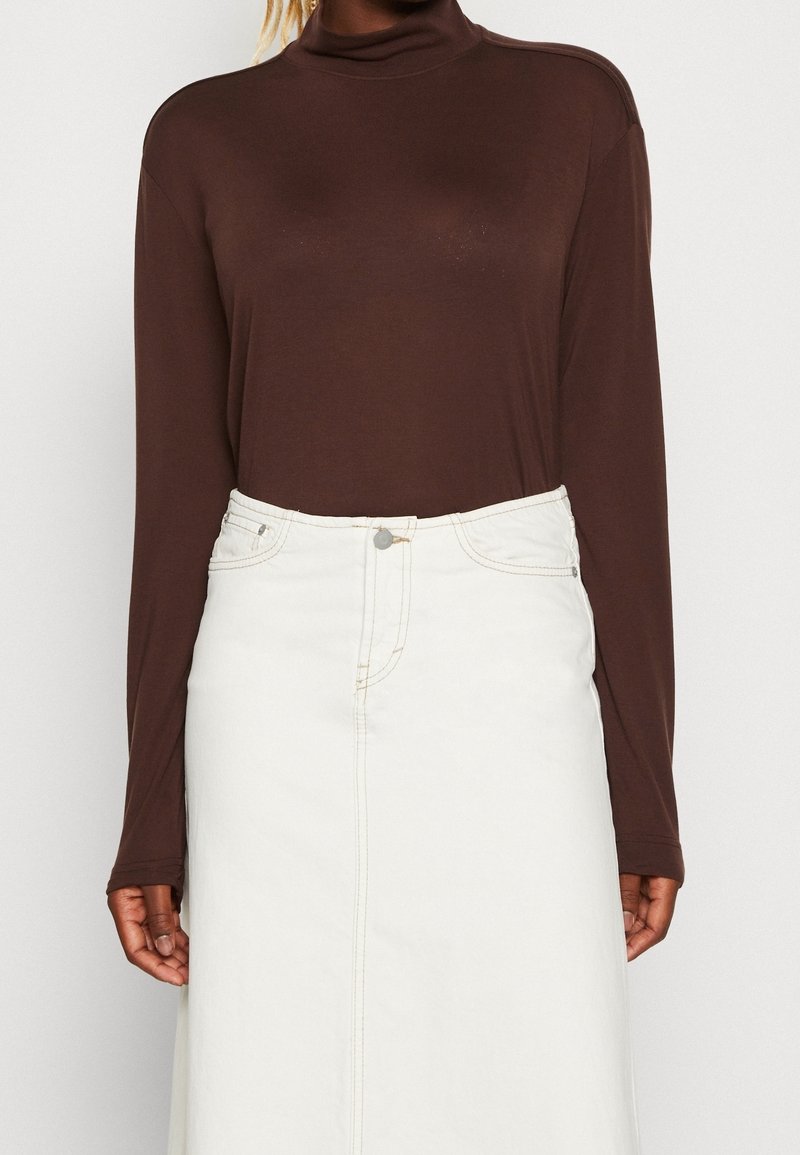 Brun långärmad topp med hög hals som är kombinerad med en vit denimkjol, som har synlig sömnad och en framknäppning med knappar.