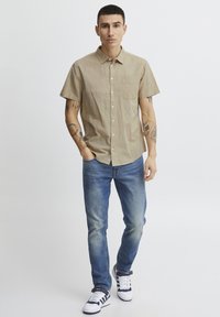 Camisa de manga corta marrón claro con un bolsillo frontal, combinada con unos vaqueros azules y zapatillas blancas con rayas marinas. Diseño simple y casual.