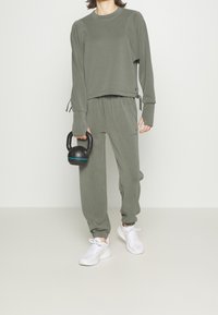 Completo sportivo verde oliva composto da top crop a manica lunga con lacci laterali e pantaloni jogger abbinati con polsini elastici, abbinato a sneakers bianche.